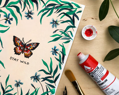 wildship studio watercolor and gouache painting of garden butterfly with green foliage and blue flowers next to a paintbrush and paint tube