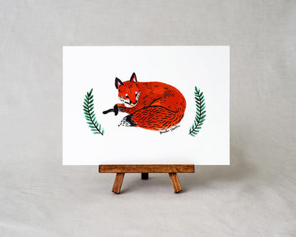 a 5x7 art print on a small easel with a watercolor painting of a red fox laying down with two branches of greenery on either side