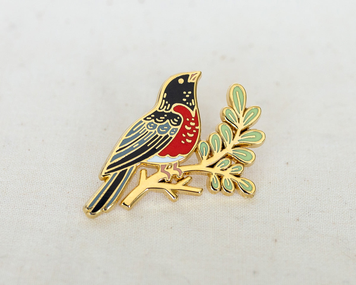 an enamel pin of a robin on a leafy branch in gold metal