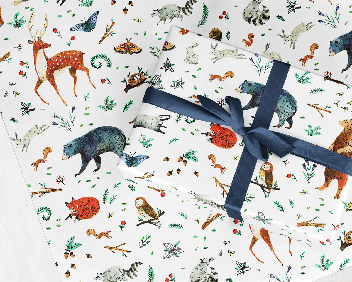 wrapped gift with a navy ribbon bow on a sheet of Wildship Studio gift wrapping paper with woodland deer, bears, raccoons, butterflies, foxes, and other animals and flowers