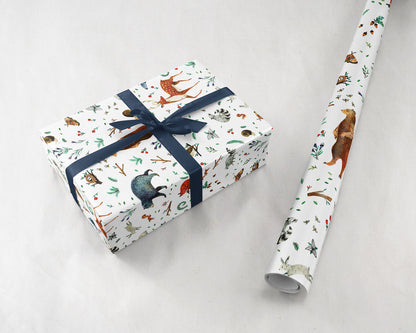 wrapped gift with a navy ribbon next to a roll of Wildship Studio gift wrapping paper with woodland deer, bears, raccoons, butterflies, foxes, and other animals and flowers