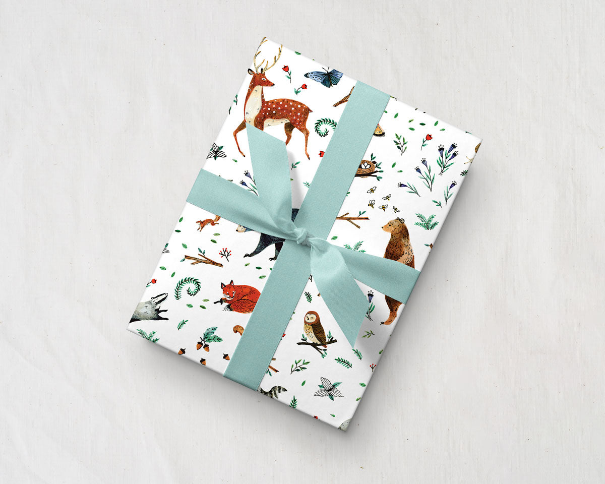 tilted gift with a blue ribbon wrapped up in Wildship Studio gift wrapping paper with woodland deer, bears, raccoons, butterflies, foxes, and other animals and flowers